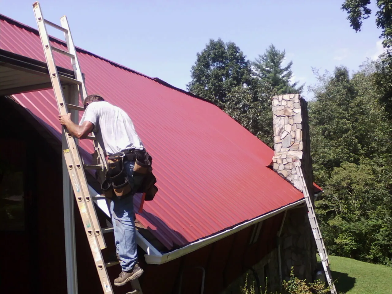 Roofer on a ladder beside a red metal roof and stone chimney — Soffit & Fascia Repair by Roofing Brothers in Tully
